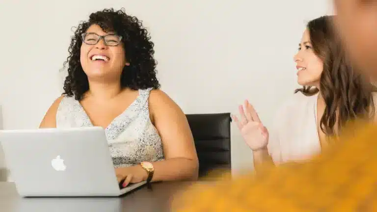 A group of women in a conference room laughing at a laptop.