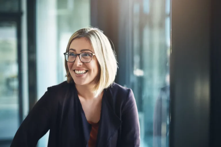 A woman wearing glasses is smiling in front of a window.